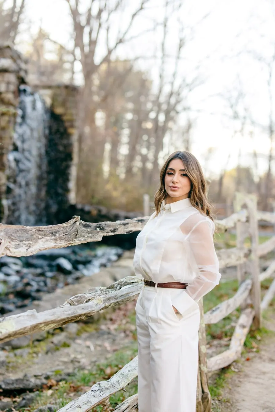 Ingrid Sampaio in white beside a rustic fence near a waterfall